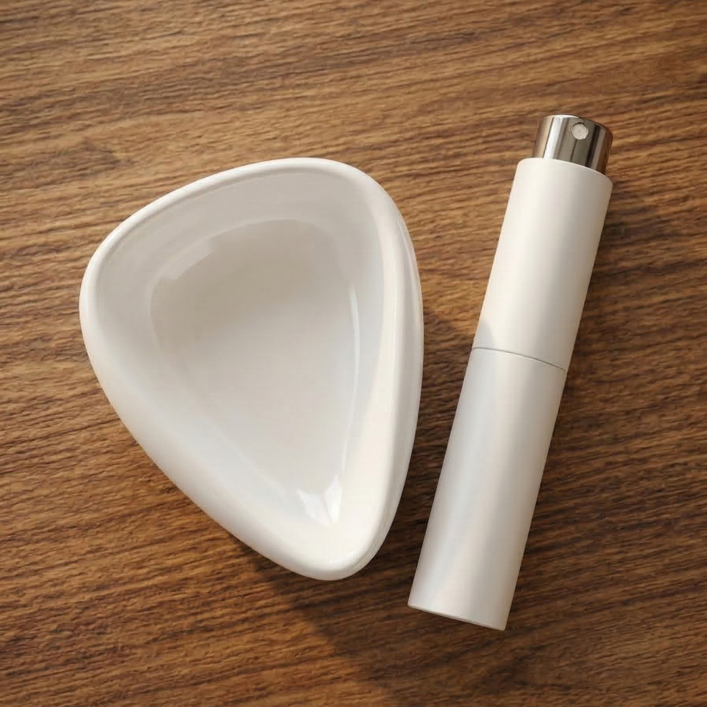 A top-down, photograph of a white ceramic coffee bean dosing tray and a matching white black RDT spray bottle resting on a warm wooden kitchen table. The lighting is soft and natural, highlighting the clean, modern textures of the barista tools.