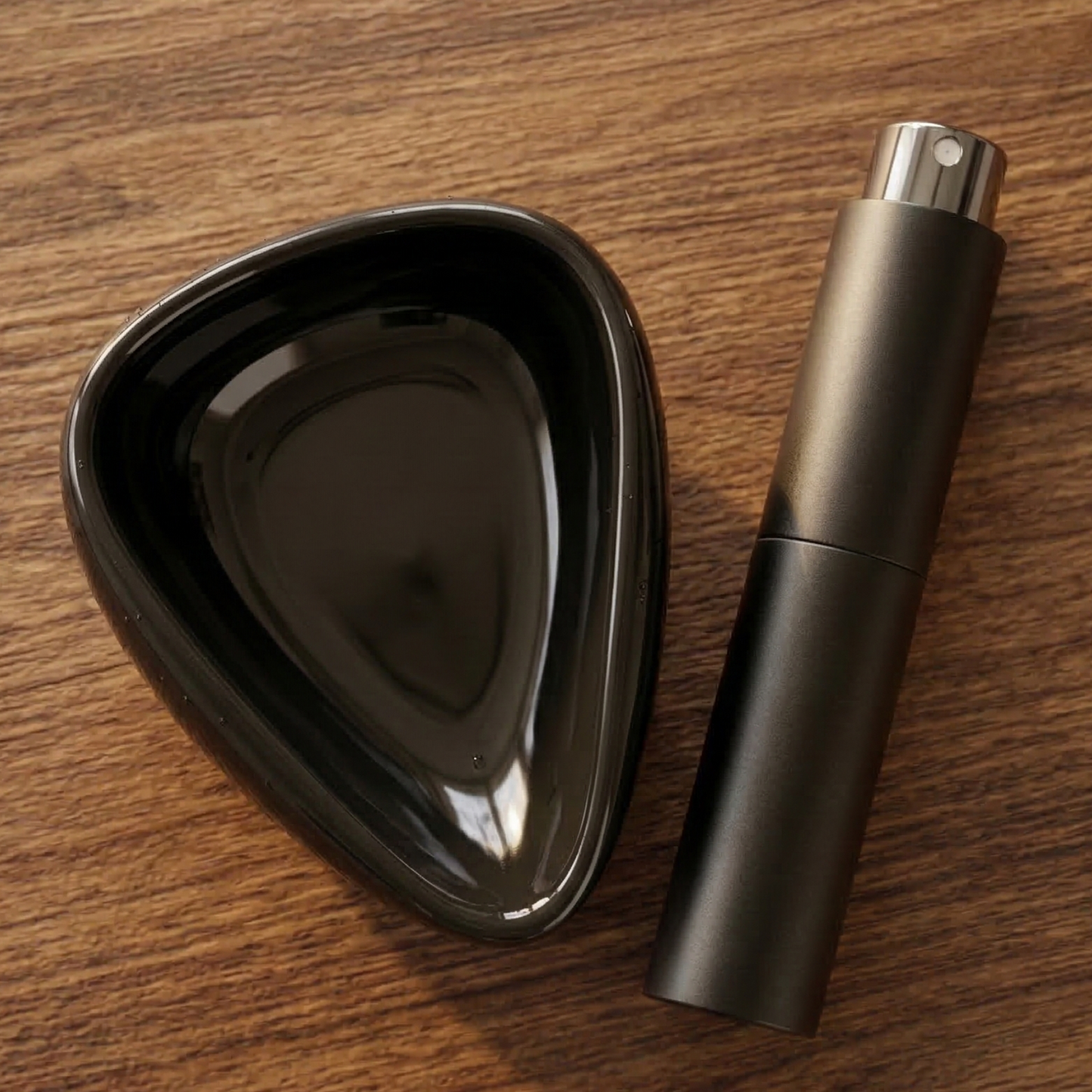 A top-down, photograph of a black ceramic coffee bean dosing tray and a matching matte black RDT spray bottle resting on a warm wooden kitchen table. The lighting is soft and natural, highlighting the clean, modern textures of the barista tools.