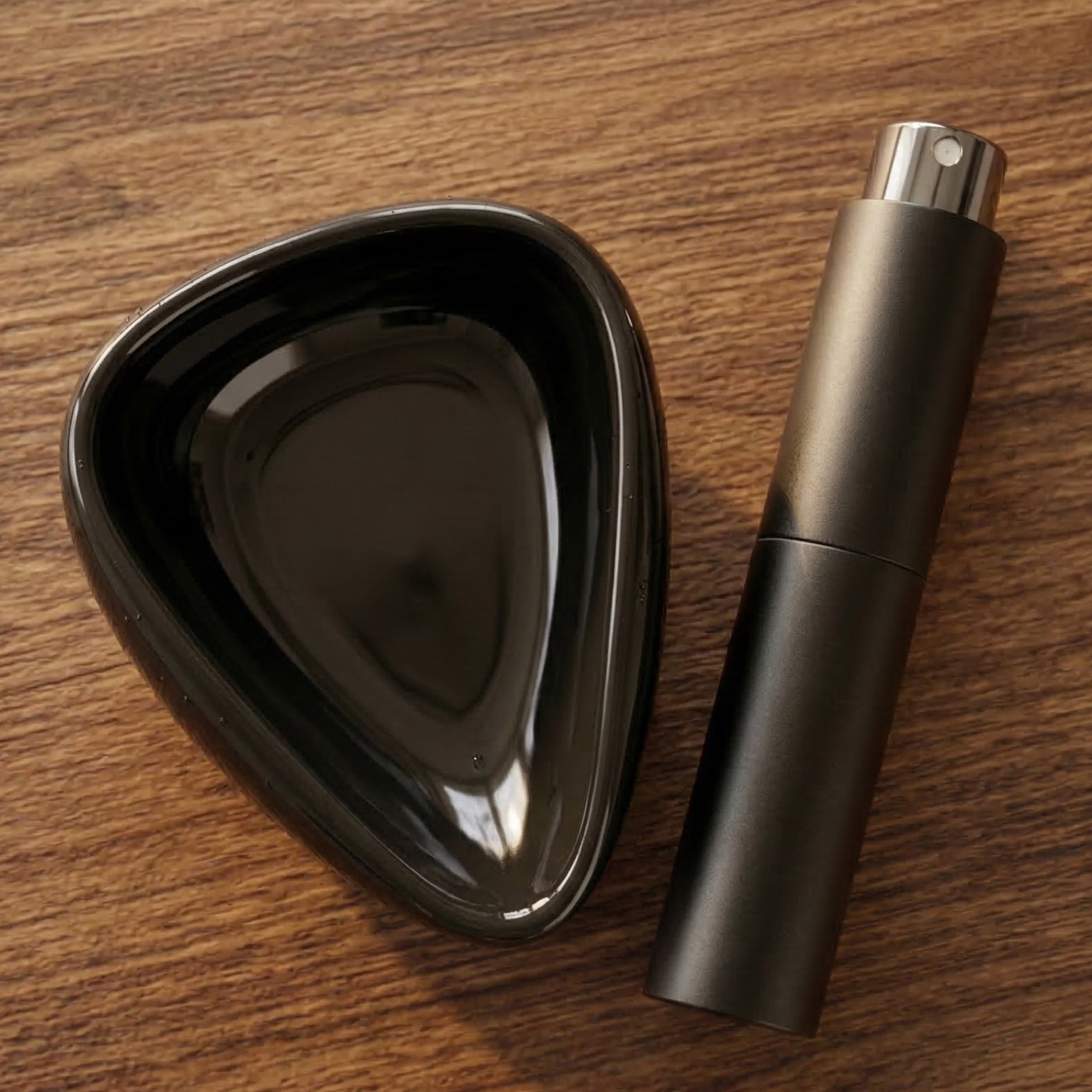 A top-down, photograph of a black ceramic coffee bean dosing tray and a matching matte black RDT spray bottle resting on a warm wooden kitchen table. The lighting is soft and natural, highlighting the clean, modern textures of the barista tools.