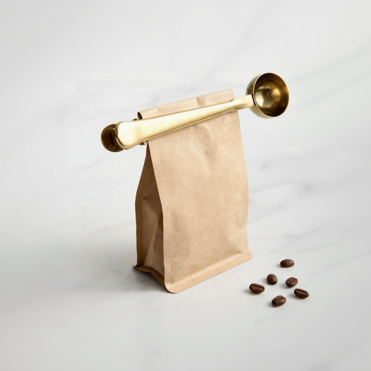 Gold coffee scoop lifting a brown paper bag with scattered coffee beans on a light background