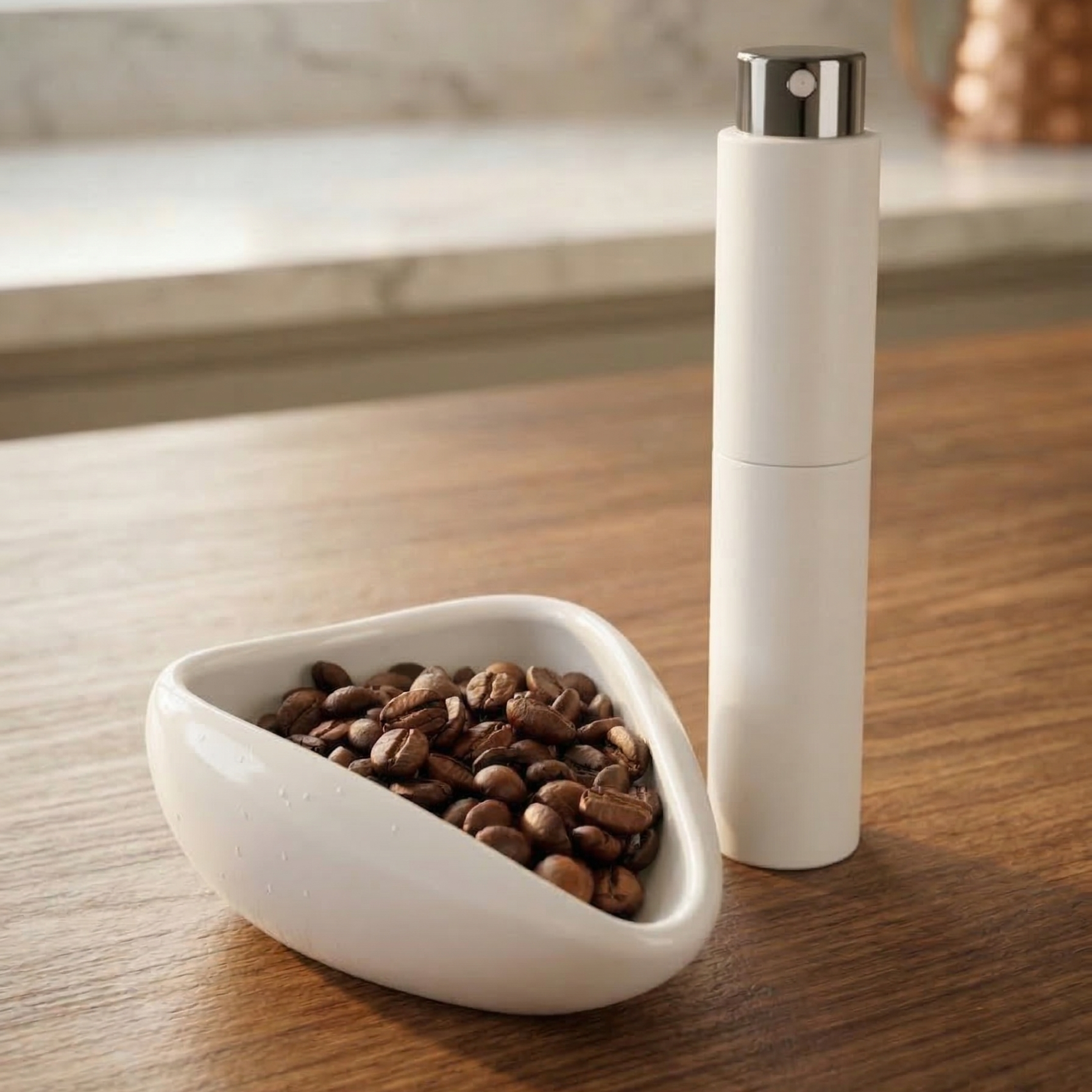 A photograph of a white ceramic coffee bean dosing tray with roasted coffee beans inside and a matching matte white RDT spray bottle resting on a warm wooden kitchen table. The lighting is soft and natural, highlighting the clean, modern textures of the barista tools.